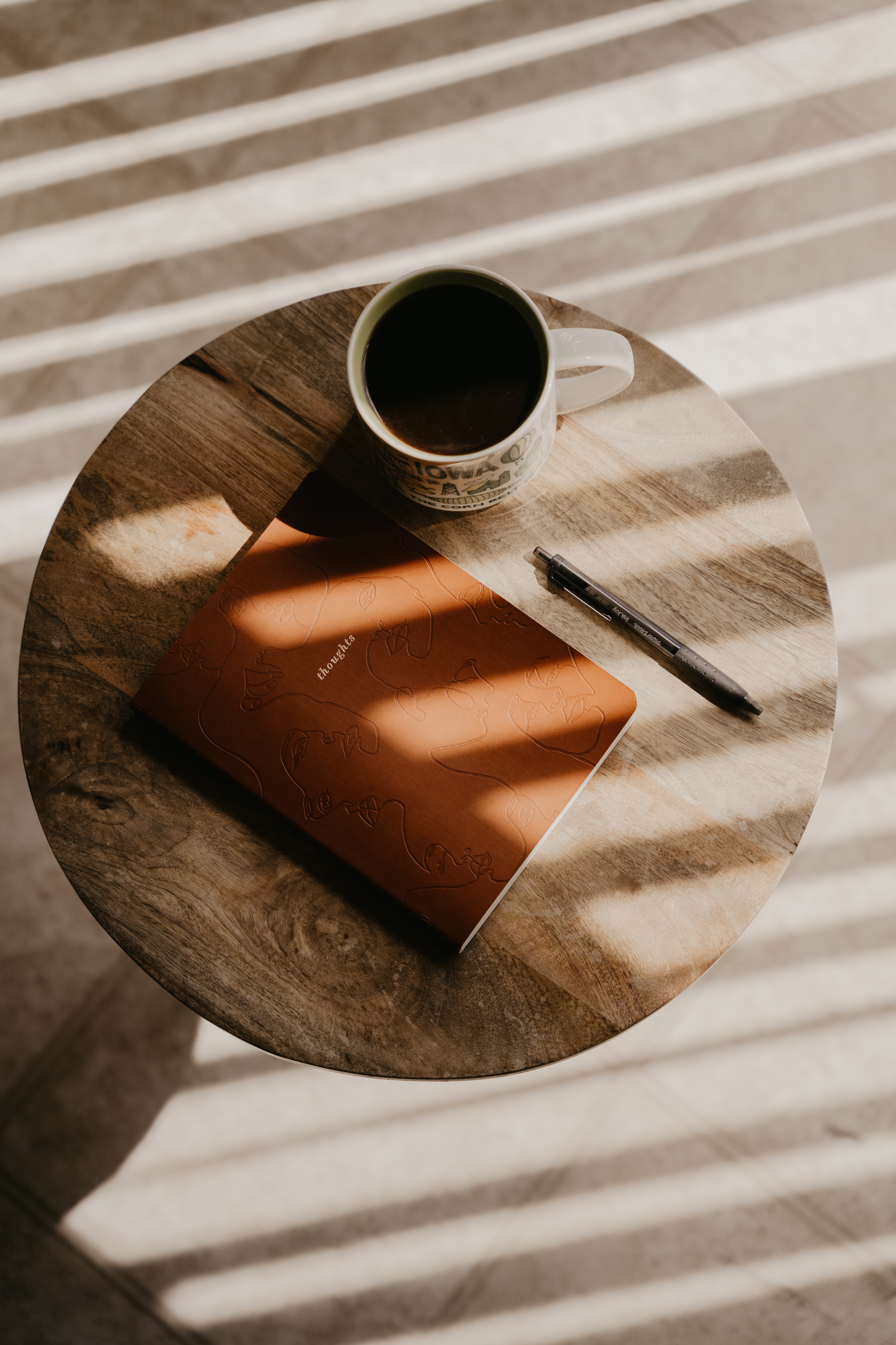 A mug of black coffee, a journal labeled “thoughts,” and a pen sit on a round wooden table with soft morning light streaming through blinds.